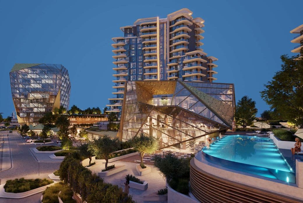 Modern urban complex with illuminated glass buildings and a rooftop infinity pool, surrounded by landscaped pathways and trees at dusk.