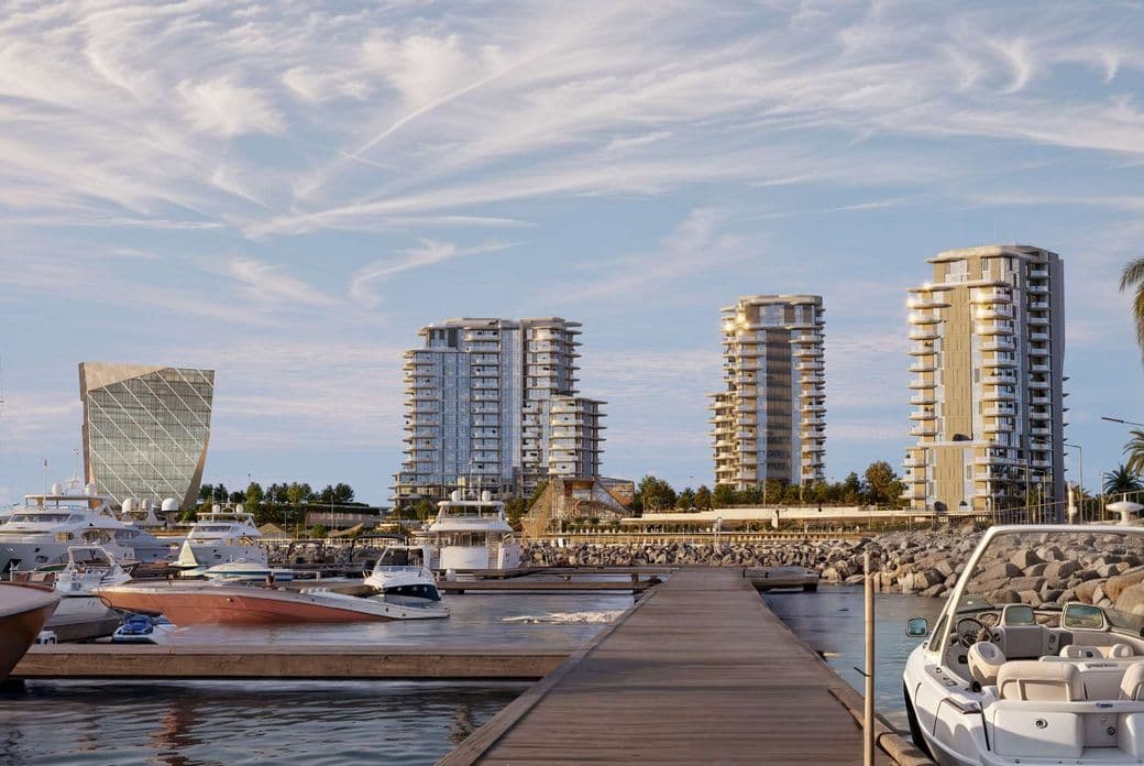 Marina with yachts, a dock, and modern high-rise buildings under a partly cloudy sky. Palm trees line the waterfront.