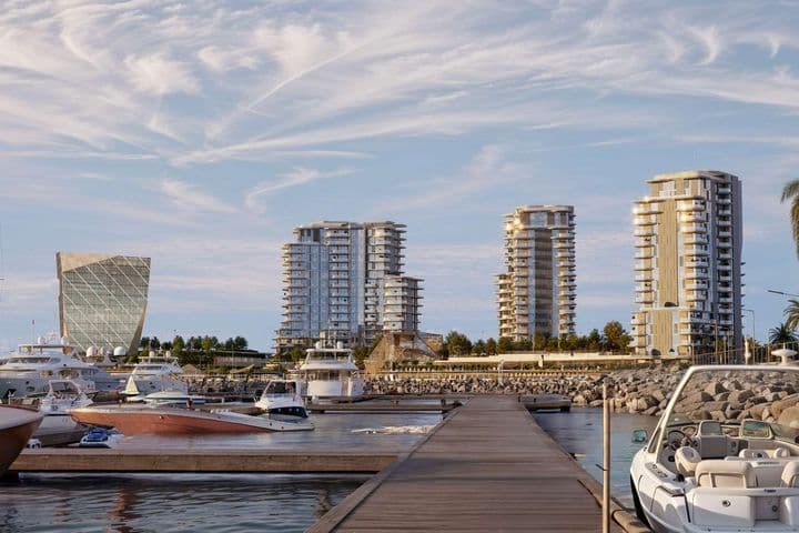 Marina with yachts, a dock, and modern high-rise buildings under a partly cloudy sky. Palm trees line the waterfront.