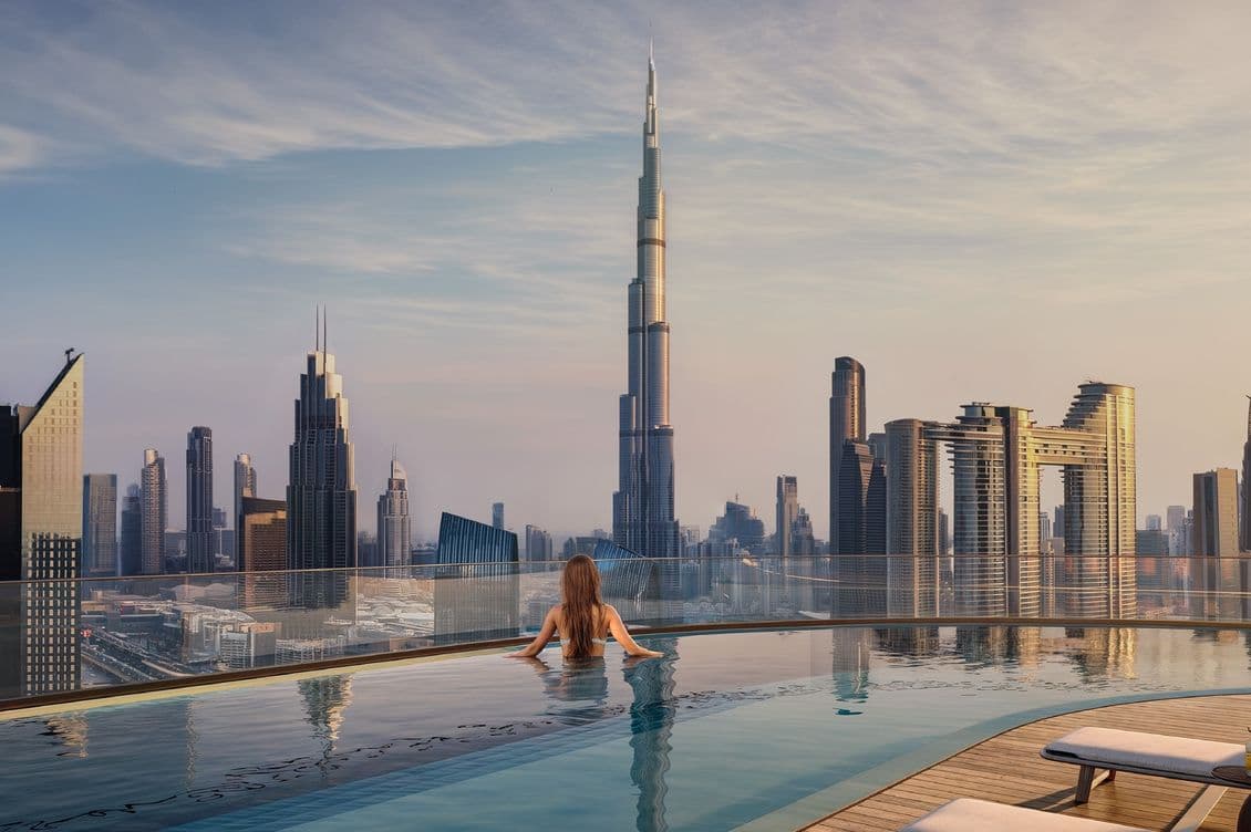 Person in rooftop pool overlooking a city skyline with a tall skyscraper at sunset, featuring modern high-rise buildings and lounge chairs.