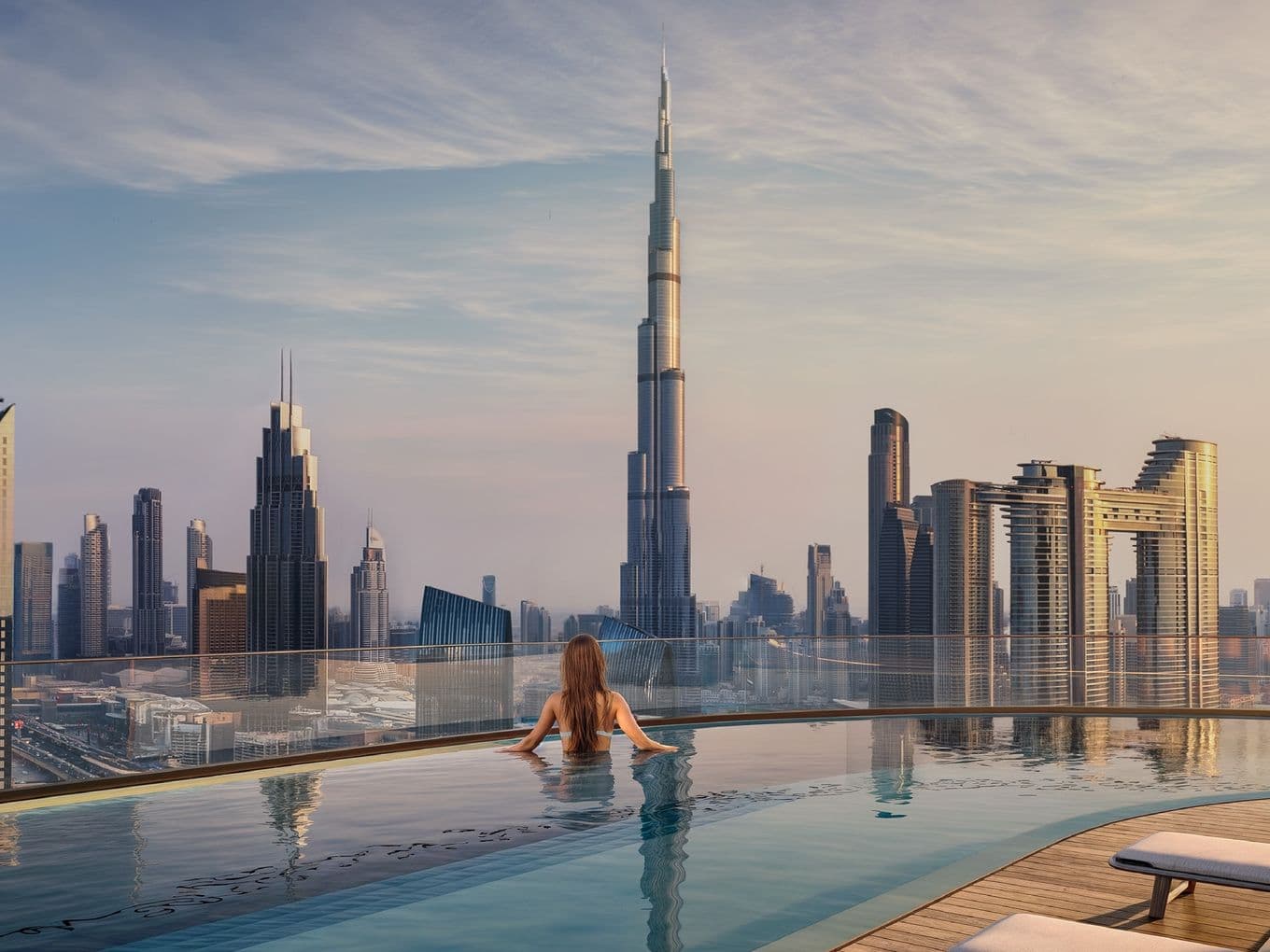 Person in rooftop pool overlooking a city skyline with a tall skyscraper at sunset, featuring modern high-rise buildings and lounge chairs.