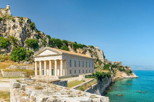 Η Παλαιά Φρουρά της Κέρκυρας παρουσιάζει ένα νεοκλασικό κτίριο με κολώνες, κοντά σε τιρκουάζ νερά και έναν βραχώδη γκρεμό με έναν φάρο.