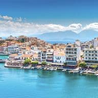 Scenic view of Agios Nikolaos, Crete, with blue water, boats, and buildings under a sunny sky. Mountains are visible in the background.