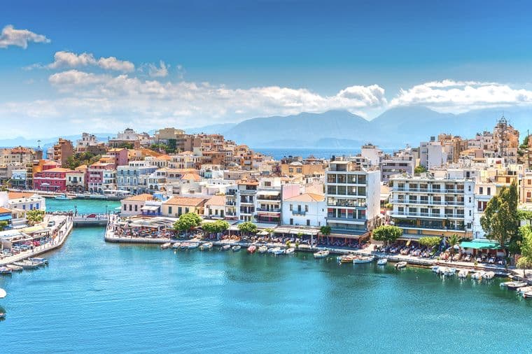 Scenic view of Agios Nikolaos, Crete, with blue water, boats, and buildings under a sunny sky. Mountains are visible in the background.