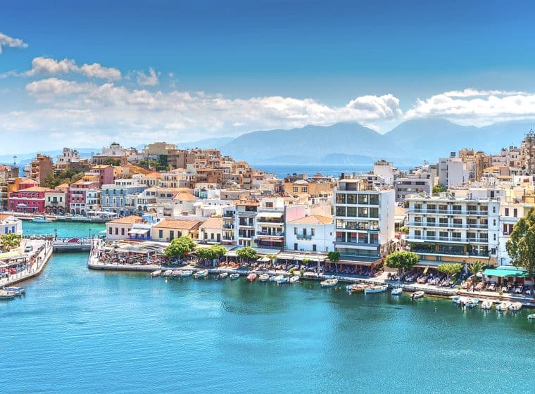 Scenic view of Agios Nikolaos, Crete, with blue water, boats, and buildings under a sunny sky. Mountains are visible in the background.