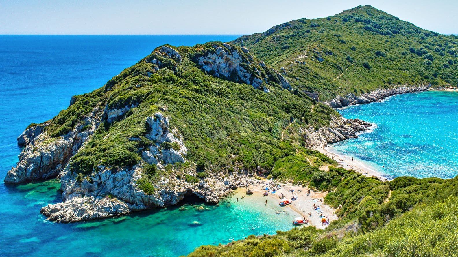 Γραφική θέα μιας καταπράσινης, βραχώδους χερσονήσου που εκτείνεται σε τιρκουάζ θάλασσα. Άνθρωποι χαλαρώνουν σε μια αμμώδη παραλία από κάτω.