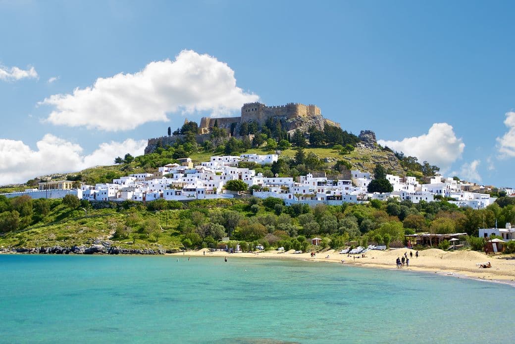 Coastal view of Lindos, Greece, with turquoise water, sandy beach, white buildings on a hillside, and a castle at the peak under a blue sky.