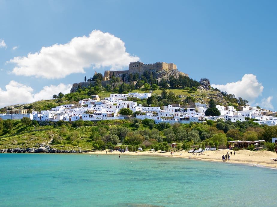 Coastal view of Lindos, Greece, with a hilltop castle overlooking white buildings, a sandy beach, and turquoise water under a blue sky.