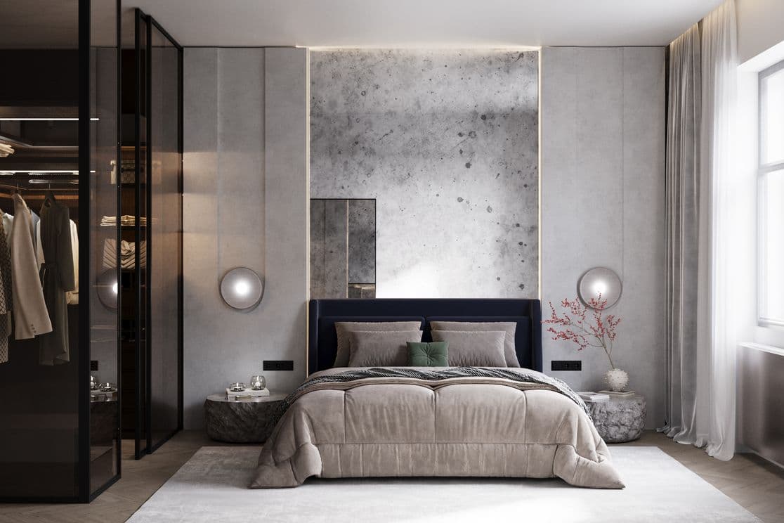 Bedroom featuring a large bed in beige tones with a dark blue headboard. Behind it is a striking concrete accent wall framed with soft indirect lighting. On both sides of the bed are round wall lamps and low stone-like bedside tables, one decorated with a vase of red branches. On the left is a glass wardrobe with dark frames, showing clothing and illuminated shelves inside. On the right, a window with long light curtains lets in natural light.