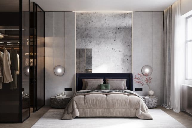 Bedroom featuring a large bed in beige tones with a dark blue headboard. Behind it is a striking concrete accent wall framed with soft indirect lighting. On both sides of the bed are round wall lamps and low stone-like bedside tables, one decorated with a vase of red branches. On the left is a glass wardrobe with dark frames, showing clothing and illuminated shelves inside. On the right, a window with long light curtains lets in natural light.