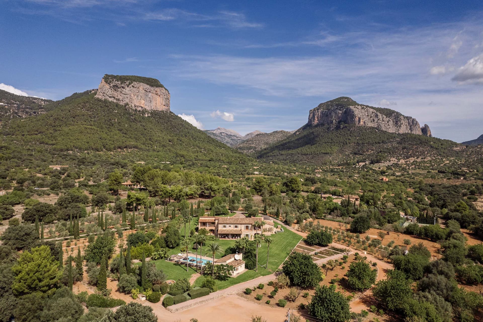 Aerial view of a Mediterranean villa surrounded by lush greenery, with mountains in the background under a clear blue sky.