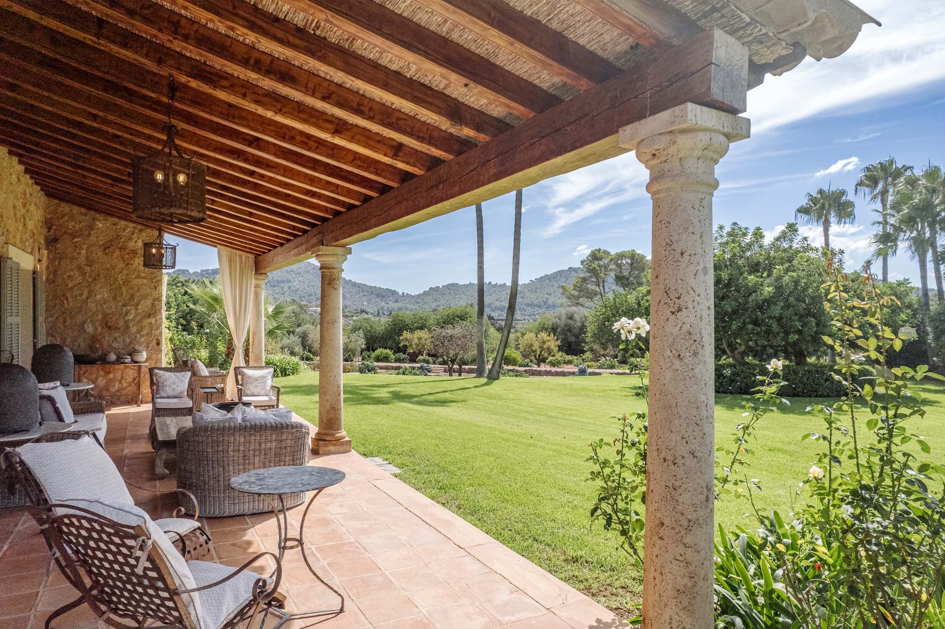 A Mediterranean-style terrace with wicker furniture overlooks the lush garden and distant hills under a clear blue sky.