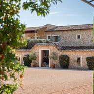 Stone house with a tiled roof, surrounded by greenery and trees, seen through an archway on a sunny day.