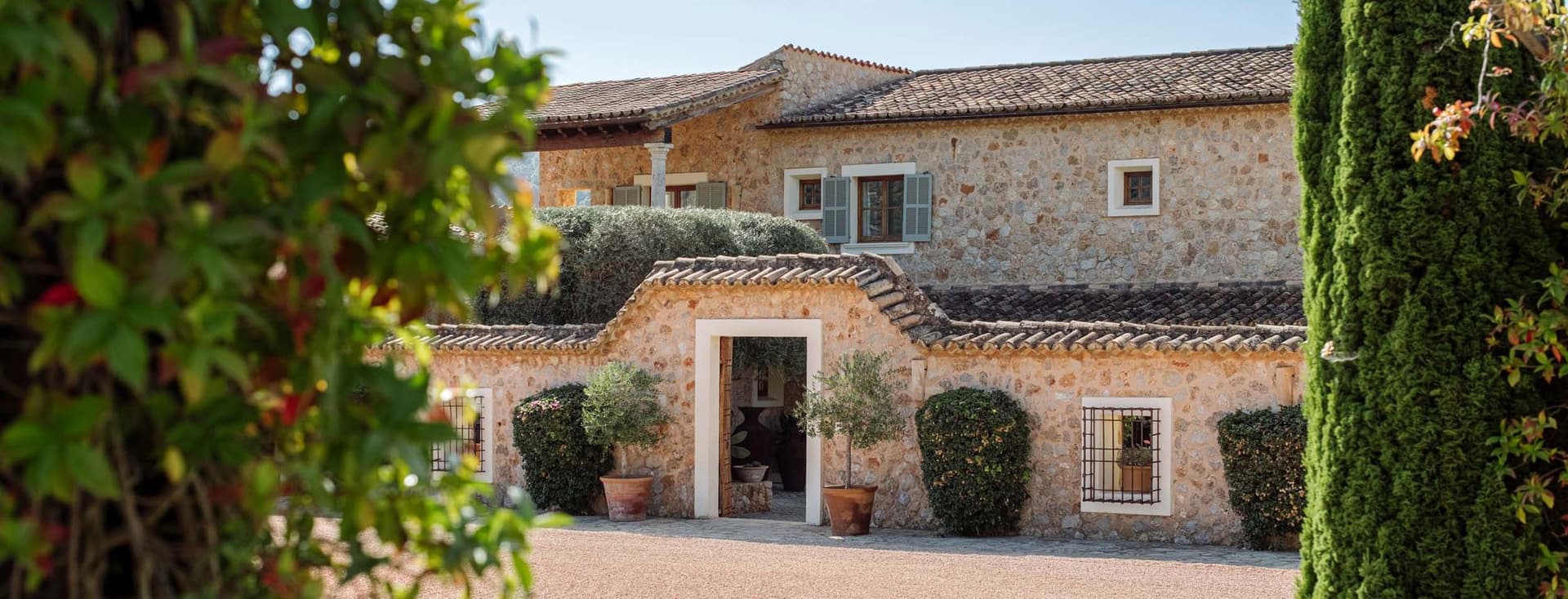 Stone house with a tiled roof, surrounded by greenery and trees, seen through an archway on a sunny day.