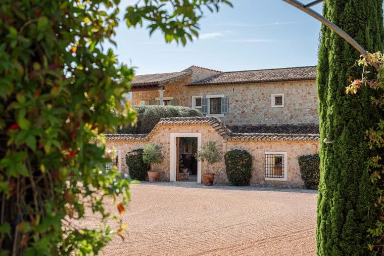 Stone house with a tiled roof, surrounded by greenery and trees, seen through an archway on a sunny day.