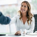 Woman in a white blouse smiling and shaking hands across a desk, with a notebook and glass of water nearby.
