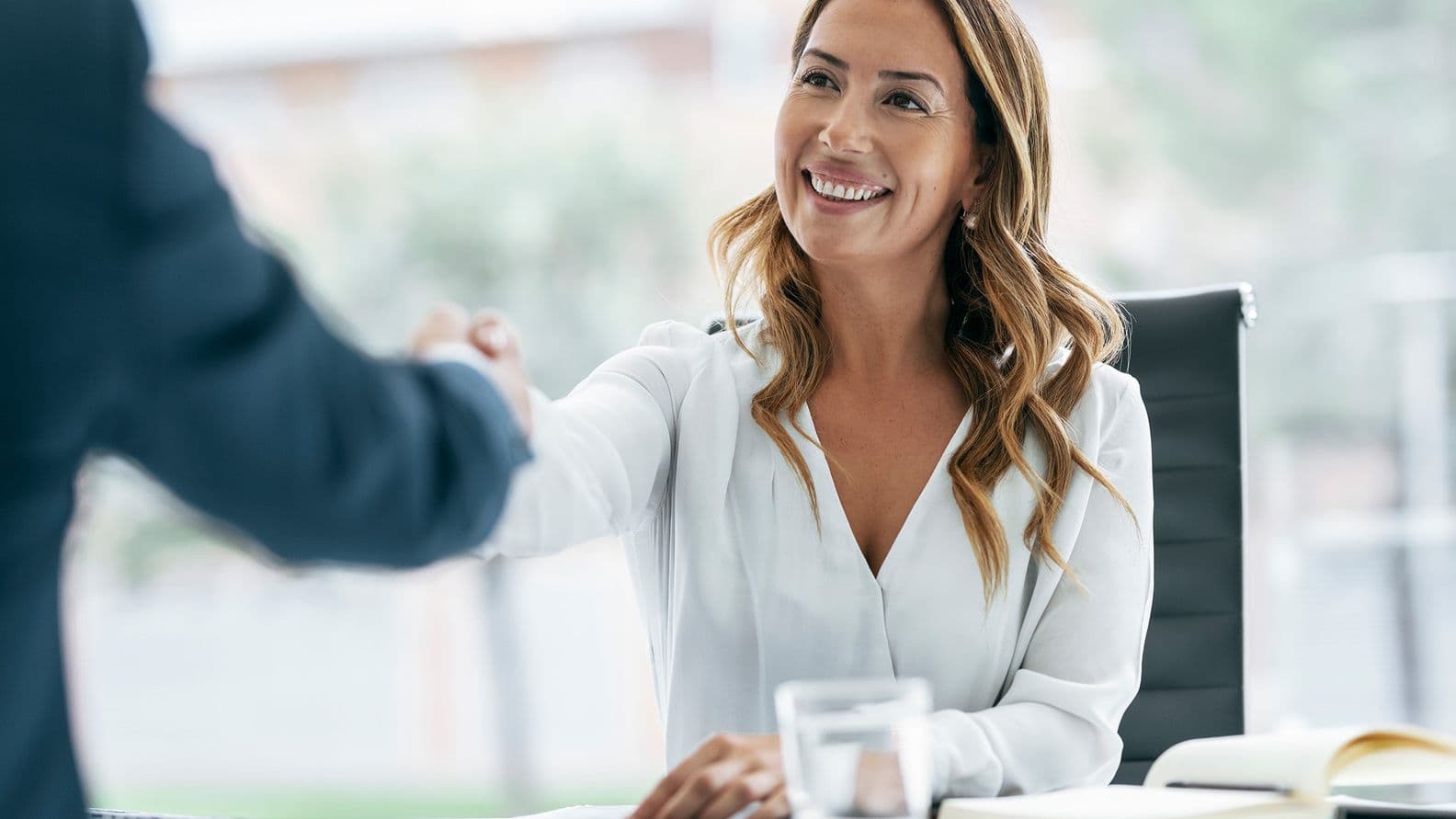 Woman in a white blouse smiling and shaking hands across a desk, with a notebook and glass of water nearby.