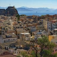 Εναέρια άποψη μιας παραθαλάσσιας πόλης με ένα μείγμα από μπεζ, κίτρινα και λευκά κτίρια κάτω από έναν καταγάλανο ουρανό. Ένα ιστορικό φρούριο είναι εμφανές στο βάθος και τα βουνά είναι ορατά απέναντι από τη θάλασσα.