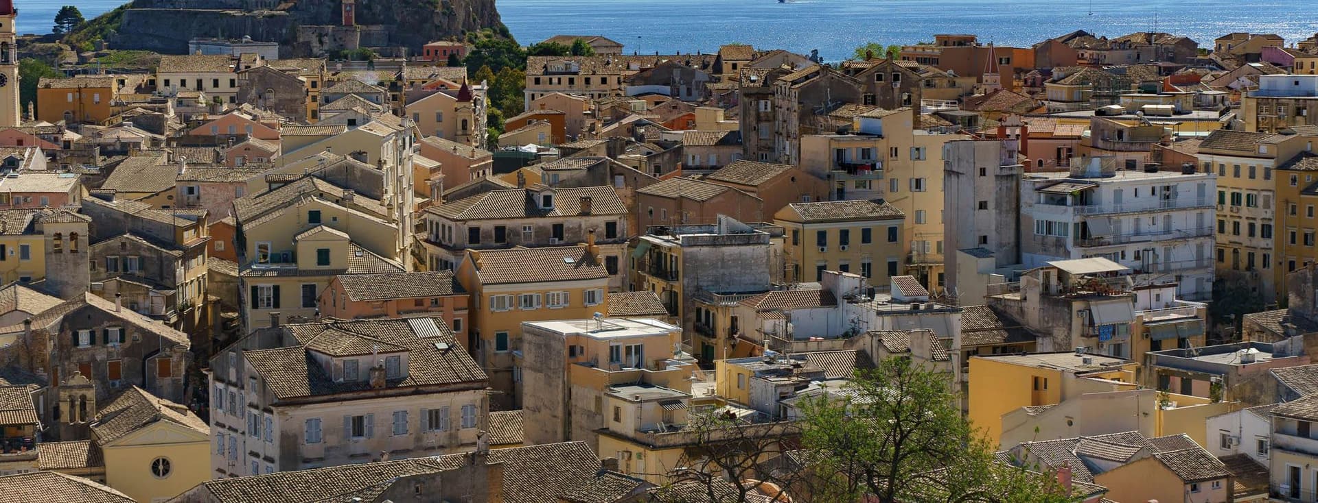 Εναέρια άποψη μιας παραθαλάσσιας πόλης με ένα μείγμα από μπεζ, κίτρινα και λευκά κτίρια κάτω από έναν καταγάλανο ουρανό. Ένα ιστορικό φρούριο είναι εμφανές στο βάθος και τα βουνά είναι ορατά απέναντι από τη θάλασσα.