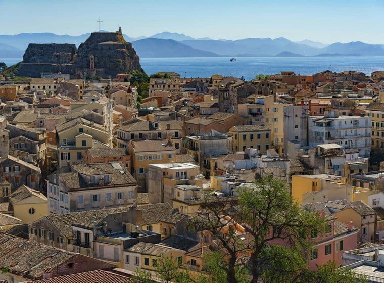 Εναέρια άποψη μιας παραθαλάσσιας πόλης με ένα μείγμα από μπεζ, κίτρινα και λευκά κτίρια κάτω από έναν καταγάλανο ουρανό. Ένα ιστορικό φρούριο είναι εμφανές στο βάθος και τα βουνά είναι ορατά απέναντι από τη θάλασσα.