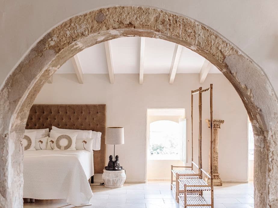 Un dormitori serè en una propietat d'estil mediterrani a Sencelles, Mallorca, amb tons neutres, un arc de pedra, sostre amb bigues i decoració rústica.