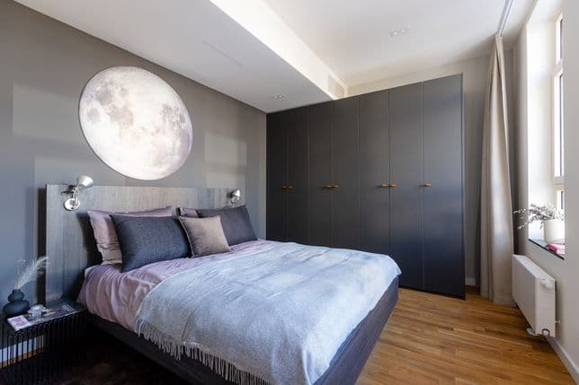 King-size bed with pastel purple bed linen, blue bedspread, a moon lamp above the bed and a dark gray wardrobe in the background. Window light comes from the right.