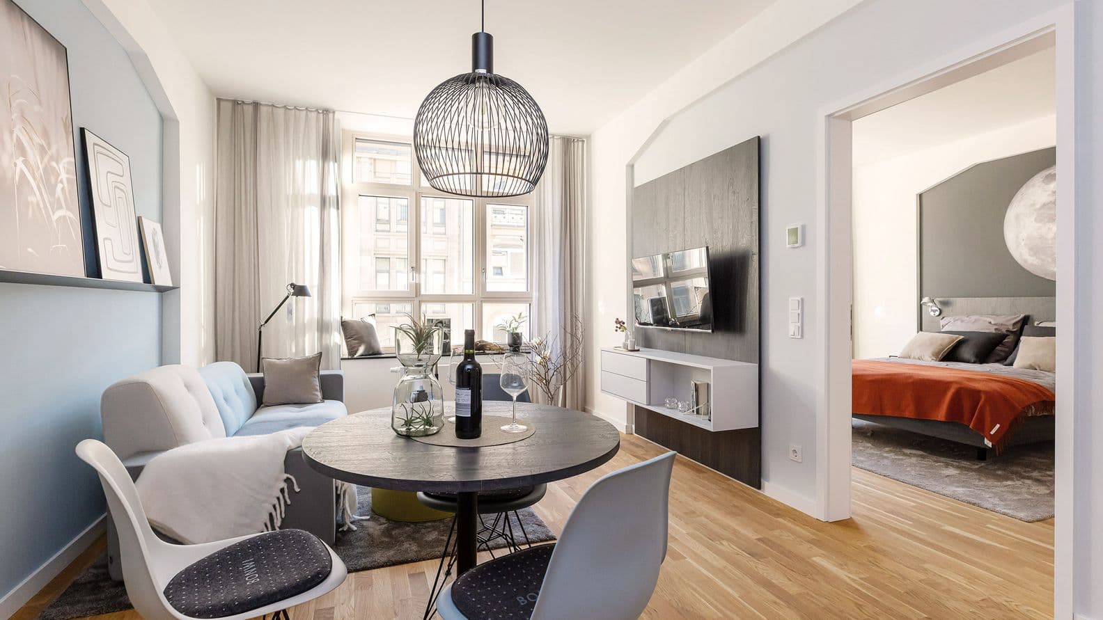 Living/dining area with two chairs and a round table in the foreground, a couch and TV corner by the window in the background. On the right, a wide white door frame without a double door leads into the bedroom.