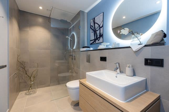 Bathroom with walk-in rain shower, separated by a fixed glass wall. A round, backlit mirror hangs above the washbasin.