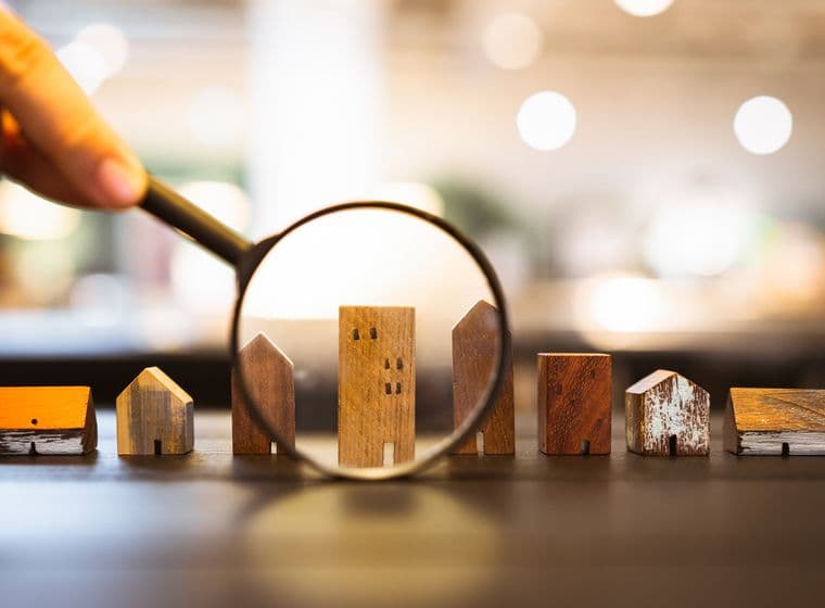 A collection of small, handcrafted wooden houses of different sizes and shapes is being viewed through a magnifying glass. The miniature houses are made from various types of wood and show signs of aging. A larger building in the middle, resembling an apartment building, is in the focus of the magnifying glass. The blurred background shows warm bokeh light effects. The scene symbolizes the careful search and analysis in real estate selection.