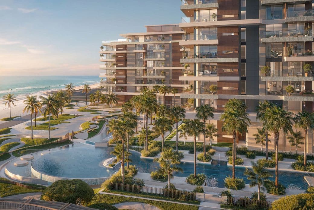 A modern beachfront residential development with glass-fronted balconies overlooking landscaped pools, palm trees, and a sandy shoreline along calm blue water at sunset.