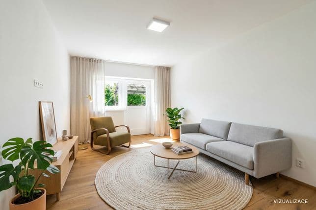 Minimalist living room with wooden flooring, a grey sofa, an olive armchair and a round rug. In the center is a low table, with a cabinet along the wall holding decor and a plant. The space is filled with natural light from a large window and a door leading to a balcony overlooking greenery.