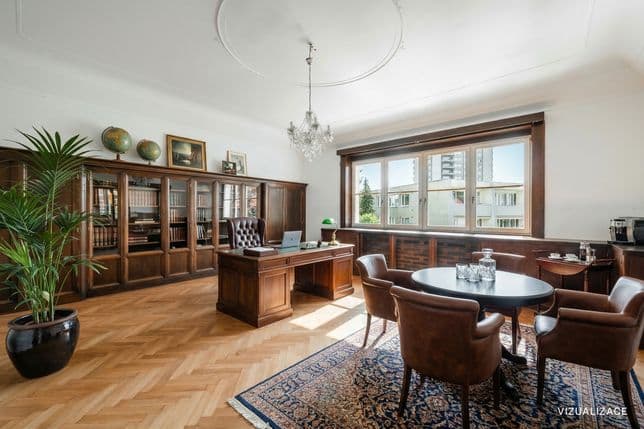 English: Spacious study with parquet flooring and solid wooden furniture. Along the wall is a bookcase with glass doors, and in the center stands a desk with a leather chair and lamp. In the foreground is a round table with four leather chairs on a patterned rug. The room is complemented by a chandelier, artwork and large windows bringing in natural daylight.