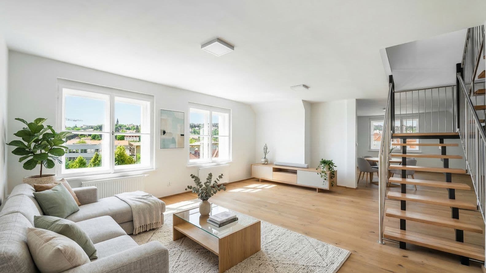 Spacious living room with wooden flooring, a corner sofa, a coffee table and a rug. Along the wall is a low cabinet with decor and plants. Large windows bring in plenty of daylight and offer views of surrounding buildings and greenery. On the right, a wooden staircase with metal railing leads to the upper floor.