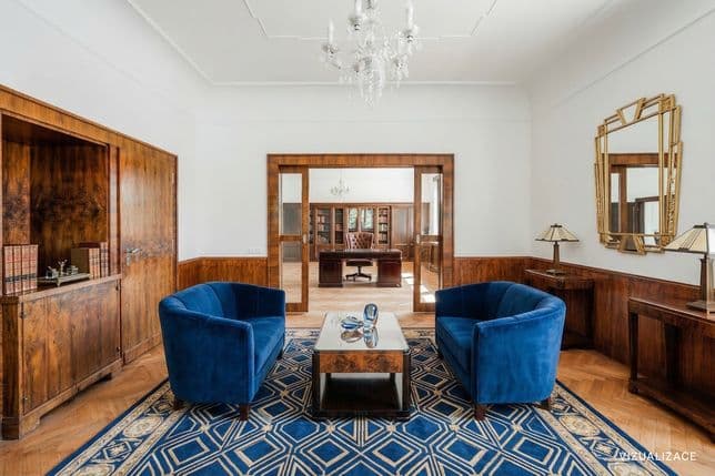 Elegant room with parquet flooring and wood-paneled walls, featuring two blue armchairs and a low table on a distinctive rug. The walls are decorated with a mirror and lamps, and a chandelier provides lighting. Through the open doors, an adjoining study with a desk and bookcase is visible.