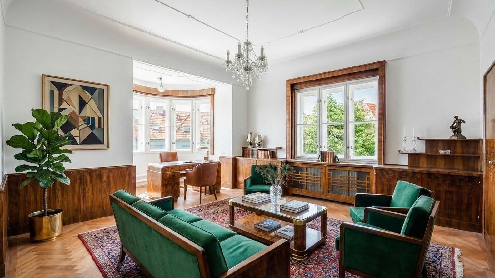 Spacious living room with parquet flooring and wood paneling, furnished with a green sofa and two armchairs around a coffee table on a patterned rug. The room is complemented by a chandelier, artwork and decor. At the back, a bay window area features a desk and views of surrounding rooftops and greenery.