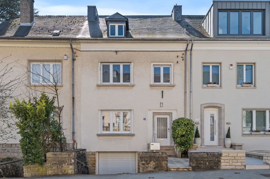 A row of three-story houses with beige exteriors, small front gardens, and a garage entrance. Clear blue sky in the background.