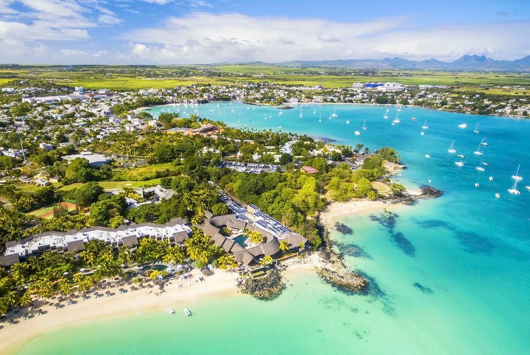Aerial view of a coastal town Grand Baie with turquoise waters, sandy beaches, boats, lush greenery, and distant mountains under a partly cloudy sky.