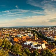 Sonnenuntergangspanorama von Graz, Österreich, mit Blick vom Schlossberg, der berühmten Uhrturm, roten Ziegeldächern, umgeben von grünen Bäumen und einer weiten Sicht auf die Stadt und die umliegenden Hügel.