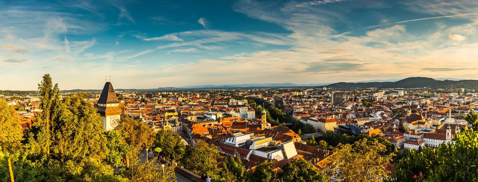 A sunset panorama of Graz, Austria, with a view from Schlossberg, featuring the famous Clock Tower, red tiled roofs, surrounded by green trees and a wide view of the city and the surrounding hills.