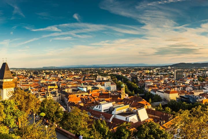 Sonnenuntergangspanorama von Graz, Österreich, mit Blick vom Schlossberg, der berühmten Uhrturm, roten Ziegeldächern, umgeben von grünen Bäumen und einer weiten Sicht auf die Stadt und die umliegenden Hügel.