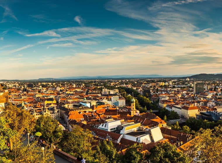 A sunset panorama of Graz, Austria, with a view from Schlossberg, featuring the famous Clock Tower, red tiled roofs, surrounded by green trees and a wide view of the city and the surrounding hills.