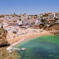 This stunning aerial view captures a picturesque Mediterranean coastal town nestled along dramatic limestone cliffs. The scene features a pristine sandy beach embraced by turquoise and emerald waters, where sunbathers dot the golden shore. Traditional white-washed buildings with terracotta roofs cascade down the hillside, creating a charming village atmosphere. The rugged coastline showcases natural rock formations and caves carved by centuries of ocean waves, while lush vegetation clings to the cliff faces. This idyllic cove represents the perfect blend of natural beauty and coastal Mediterranean architecture, offering visitors a tranquil escape with crystal-clear waters ideal for swimming and relaxation.