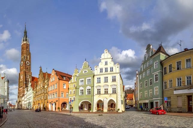 A picturesque European town square with colorful historic buildings, a tall clock tower, and a cobblestone street under a partly cloudy sky.