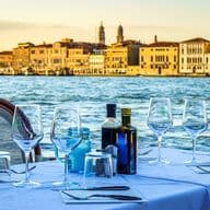 Ein Tisch ist elegant gedeckt in einem Freiluftrestaurant mit Blick auf den Canal Grande in Venedig, mit Weingläsern, Flaschen mit Öl und Essig und Besteck auf einer weißen Tischdecke. Im Hintergrund spiegeln sich die historischen Fassaden venezianischer Gebäude im Wasser bei Sonnenuntergang und schaffen eine fesselnde Atmosphäre.