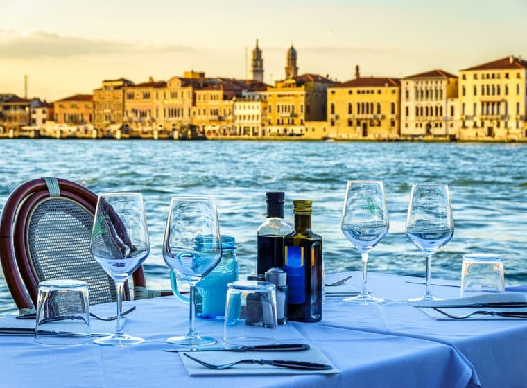 Ein Tisch ist elegant gedeckt in einem Freiluftrestaurant mit Blick auf den Canal Grande in Venedig, mit Weingläsern, Flaschen mit Öl und Essig und Besteck auf einer weißen Tischdecke. Im Hintergrund spiegeln sich die historischen Fassaden venezianischer Gebäude im Wasser bei Sonnenuntergang und schaffen eine fesselnde Atmosphäre.