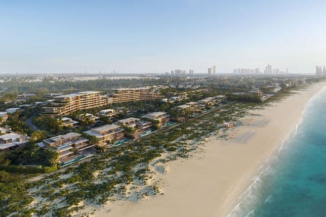 Aerial view of beachfront resort with modern villas, lush greenery, and loungers on a sandy beach beside clear blue water under a bright sky.
