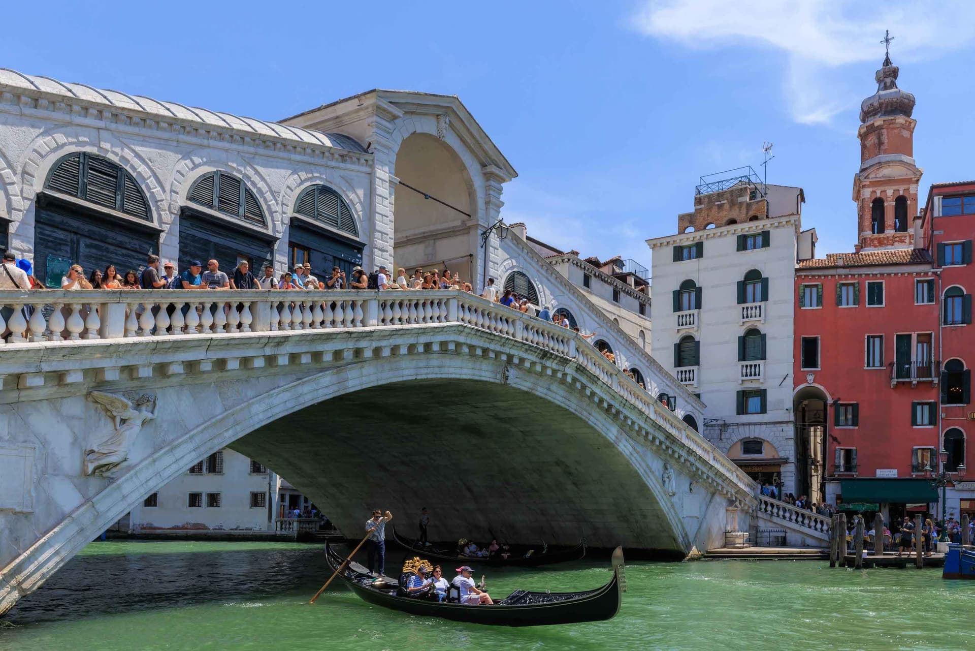 Ein Panoramablick auf die berühmte Rialtobrücke in Venedig, Italien. Die gewölbte Brücke aus weißem Stein ist oben mit Touristen bevölkert, während unten eine traditionelle Gondel mit Passagieren vorbeifährt. Die Gondel wird von einem Gondoliere in traditioneller Kleidung gesteuert. Der Canal Grande erscheint im Sonnenlicht grün, und im Hintergrund sind farbenfrohe venezianische Gebäude, darunter weiße und terrakottafarbene Bauten mit einem Glockenturm, zu sehen, alles eingerahmt von einem klaren blauen Himmel.
