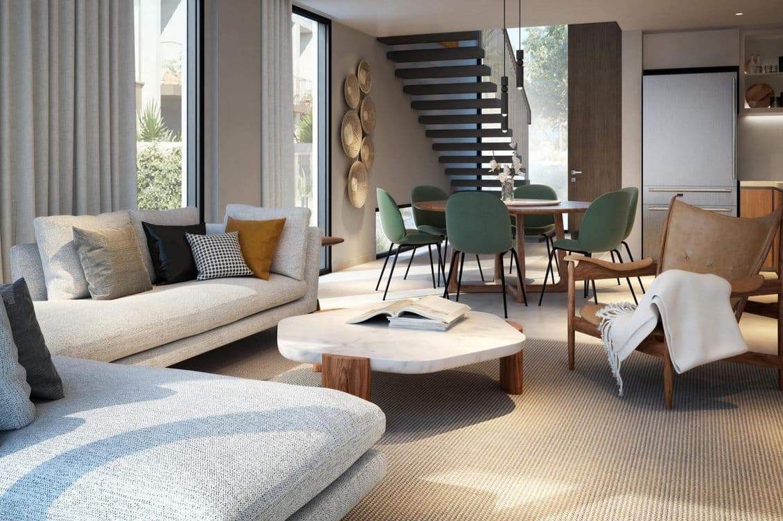 Modern living room with gray sofas, cushioned chairs, and a marble coffee table. Open dining area with green chairs and a staircase.