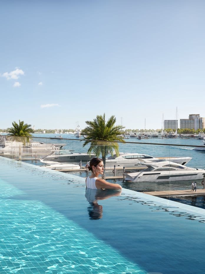 Woman relaxing in an infinity-edge pool with clear blue water, surrounded by palm trees and docked yachts, with residential towers in the background.