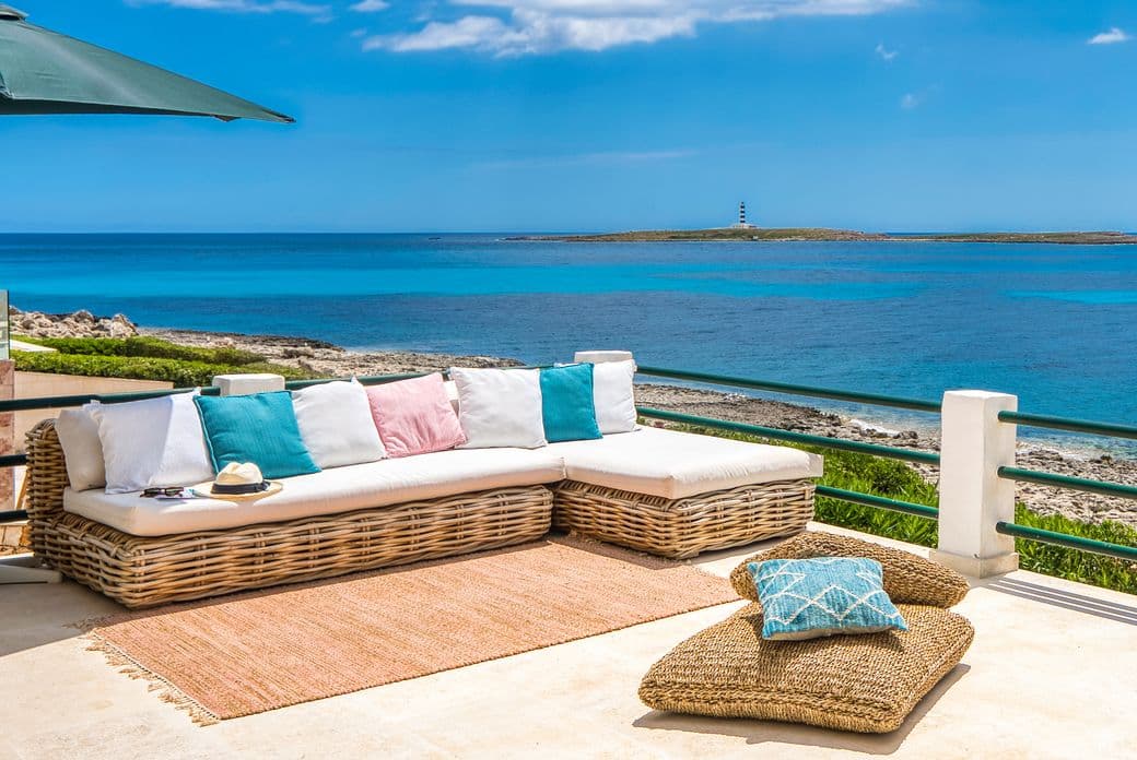 Stylish terrace with wicker furniture and colourful cushions overlooking the turquoise sea and rocky coastline in Menorca, with a distant lighthouse on the horizon.